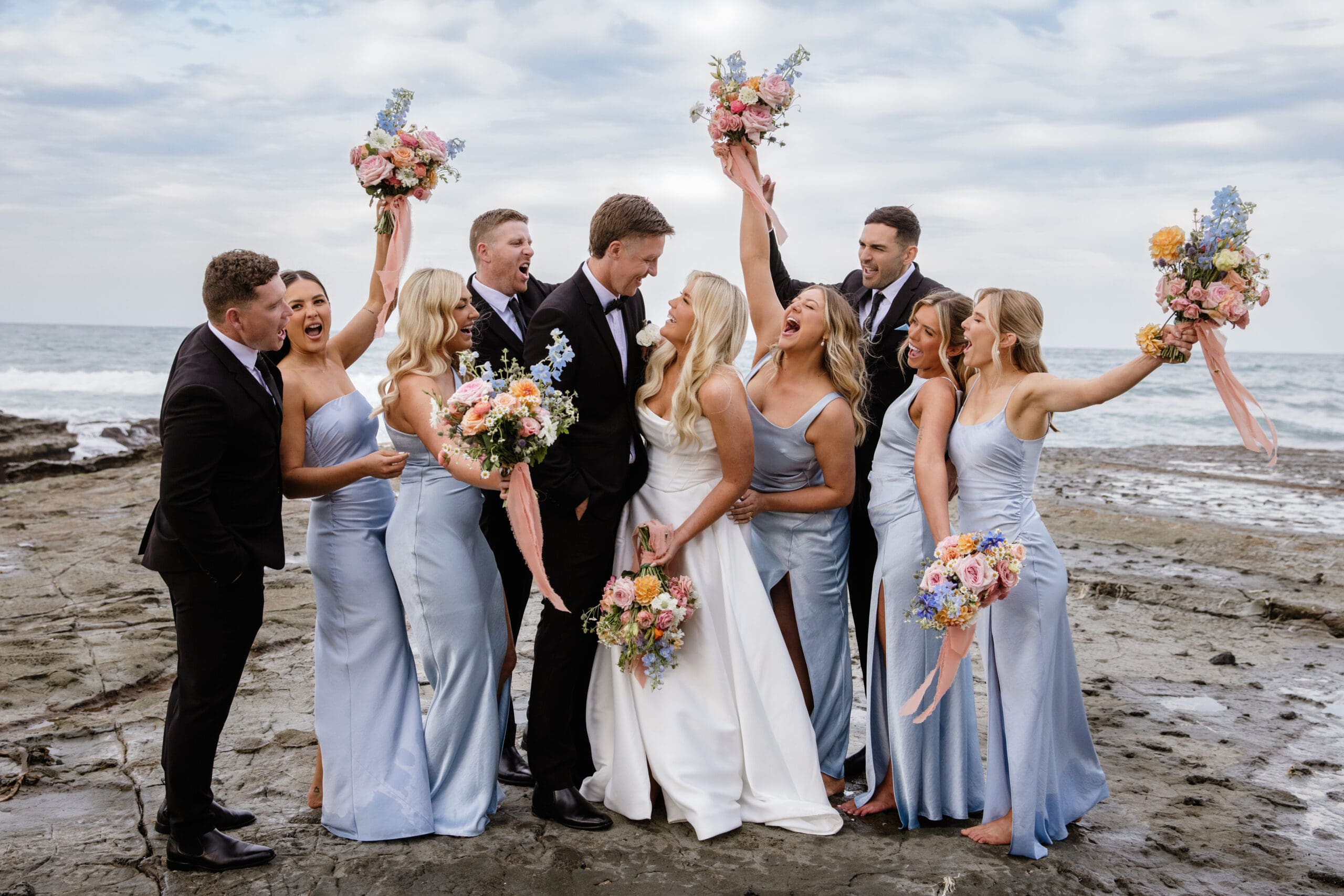 Bridal party standing on rocks by the water, cheering and raising bouquets.
