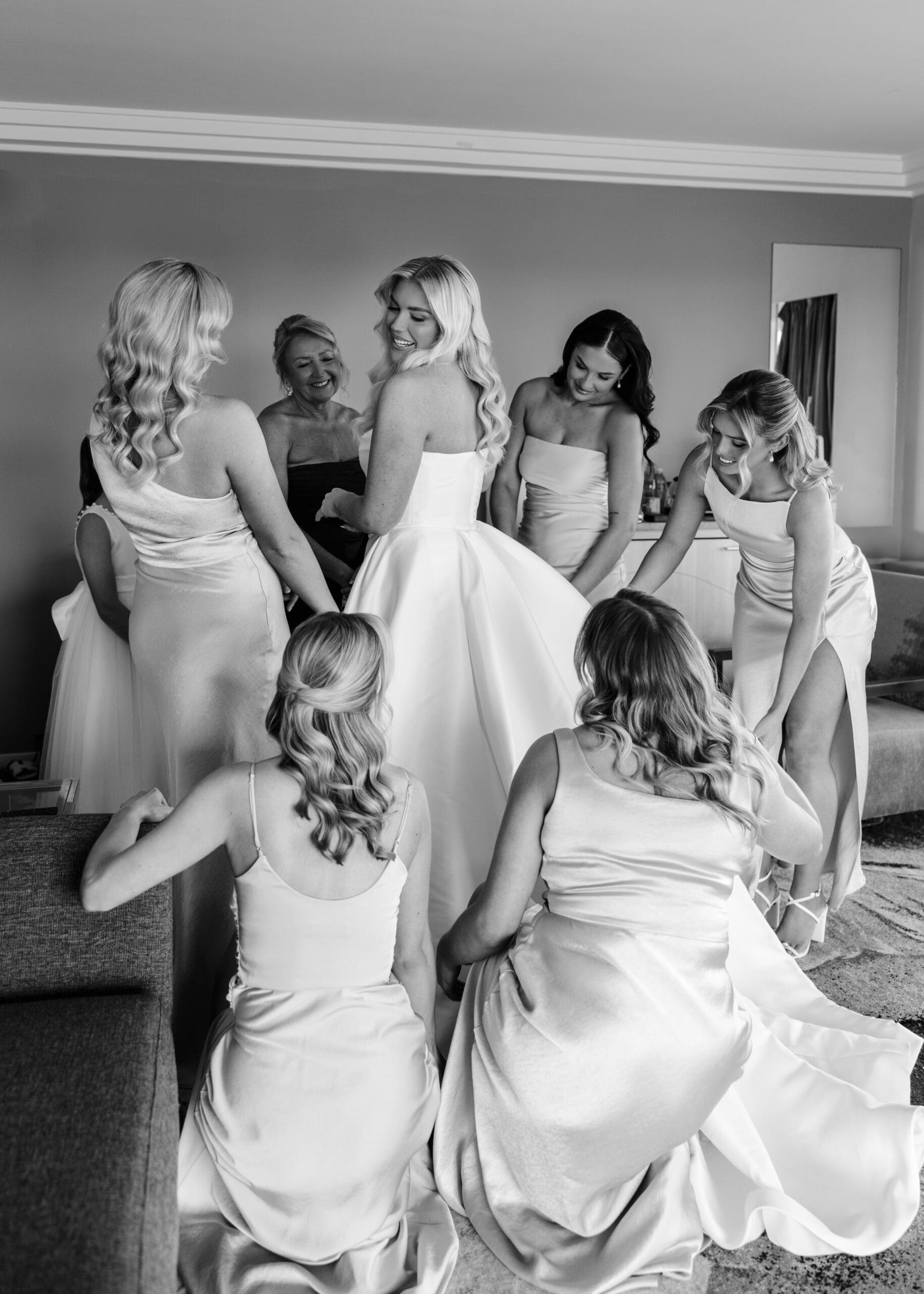 Bride in a strapless gown surrounded by bridesmaids helping adjust her dress while getting ready.