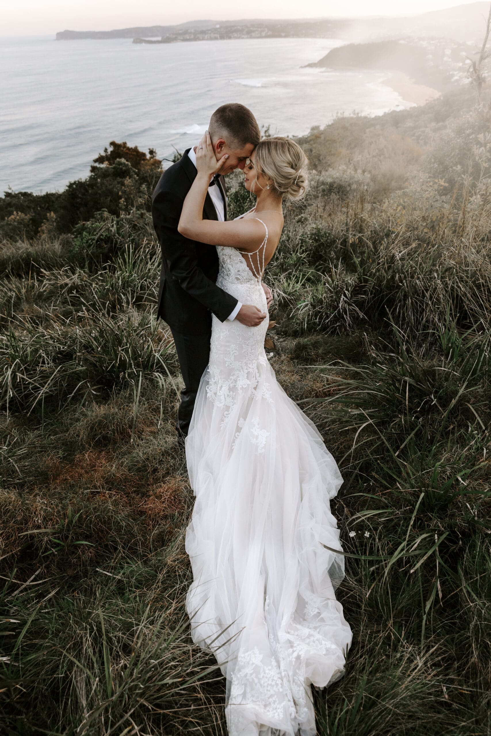Bride and groom embracing on a windy coastal cliff during wedding portraits.